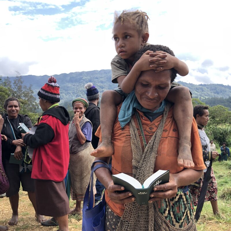A child sits on the shoulders of a man reading the Bible in Papua New Guinea.
