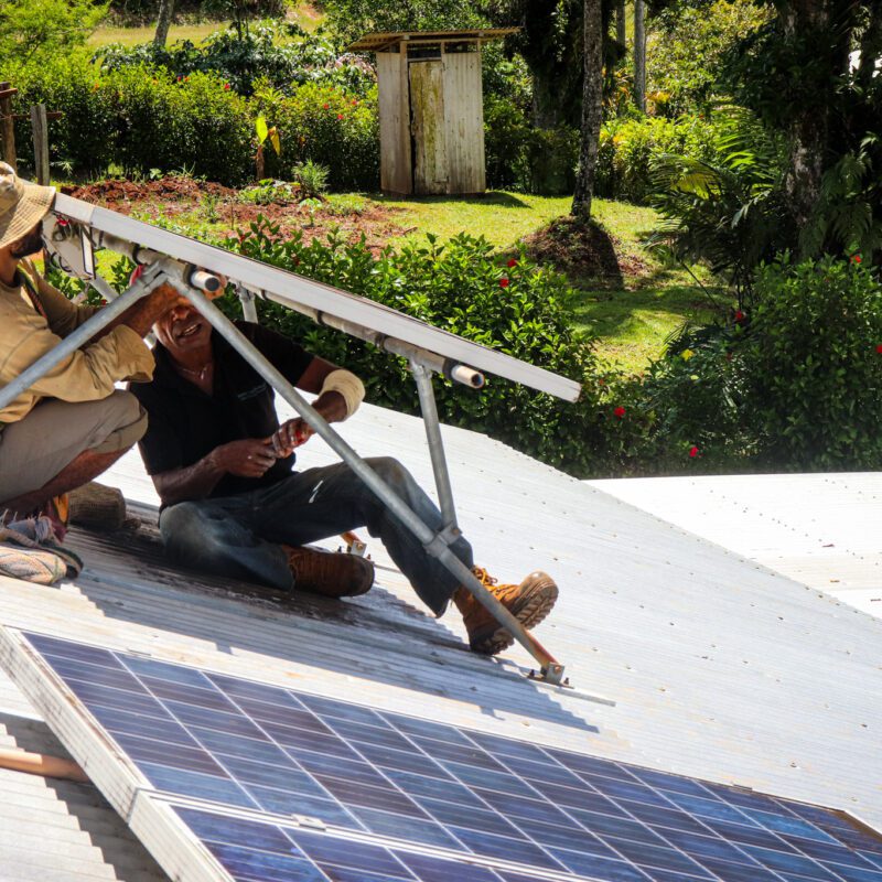 Two men sit underneath solar panels they are installing.